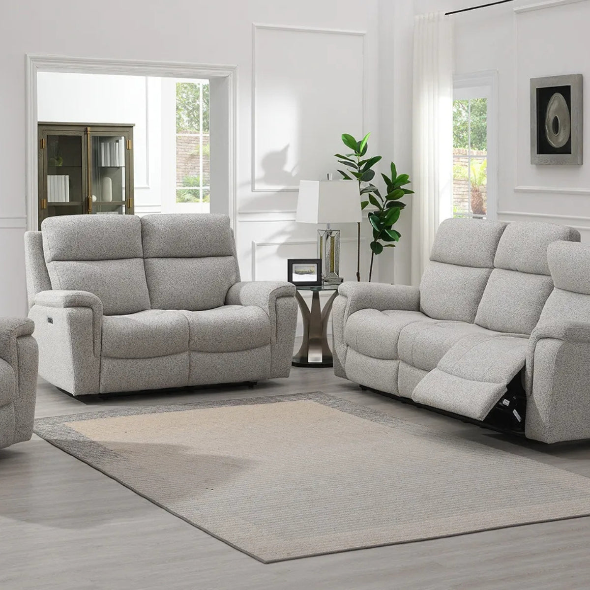 Hector grey upholstered reclining loveseat and matching reclining sofa set shown in a bright, modern living room with a beige area rug, side table, lamp, and plant.