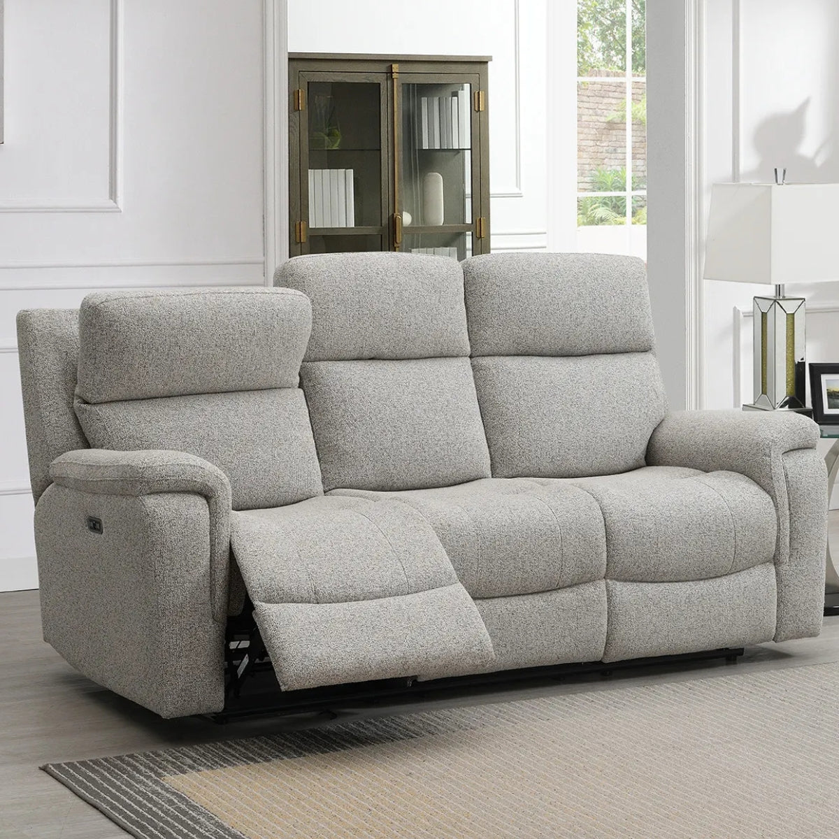 Hector grey upholstered reclining 3 seater, shown in a bright living room setting with a window, cabinet, and lamp.