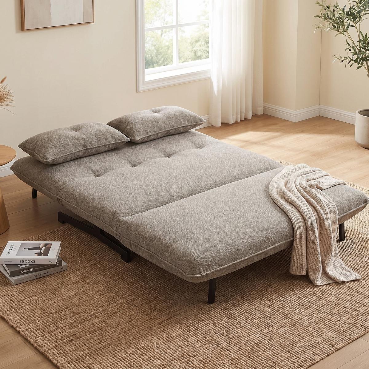 A gray fabric futon sofa bed with two matching pillows and a draped cream throw blanket, shown in a bright, modern room with a window, curtains, and a woven rug.
