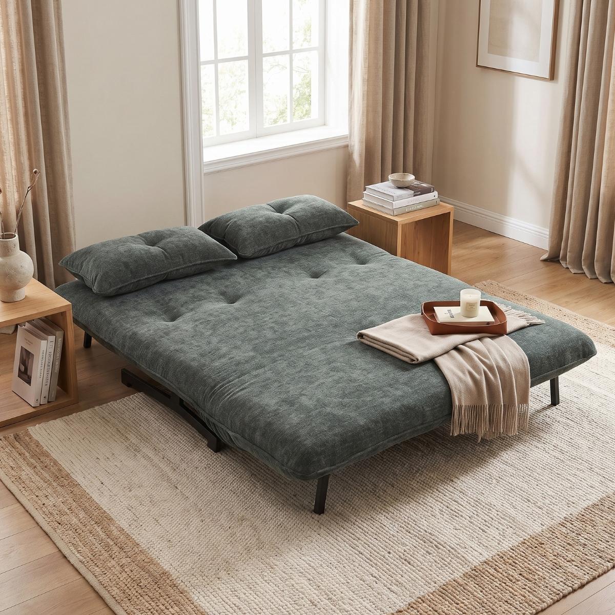 A dark green tufted futon sofa bed with two matching pillows, styled in a bright, modern bedroom with beige curtains, wooden side tables, and a striped rug.