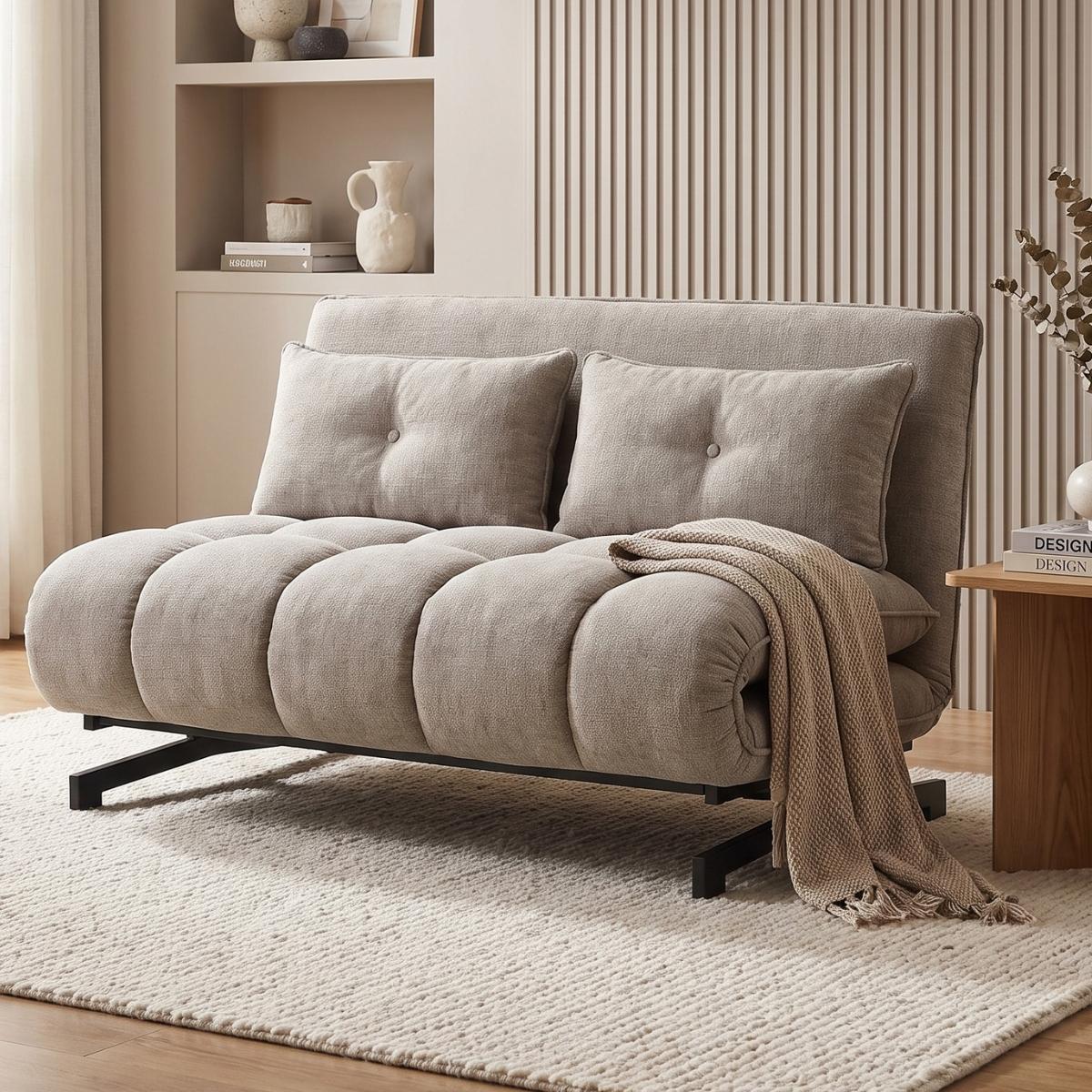 A light beige upholstered futon sofa with two matching tufted back cushions and a draped throw blanket, shown in a styled living room setting with a rug, shelving, and side table.