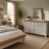 A styled bedroom scene featuring the grey Canter chest of drawers with a natural wood top and mirror, alongside a matching bed frame, in a softly lit room with sage green walls and neutral decor.