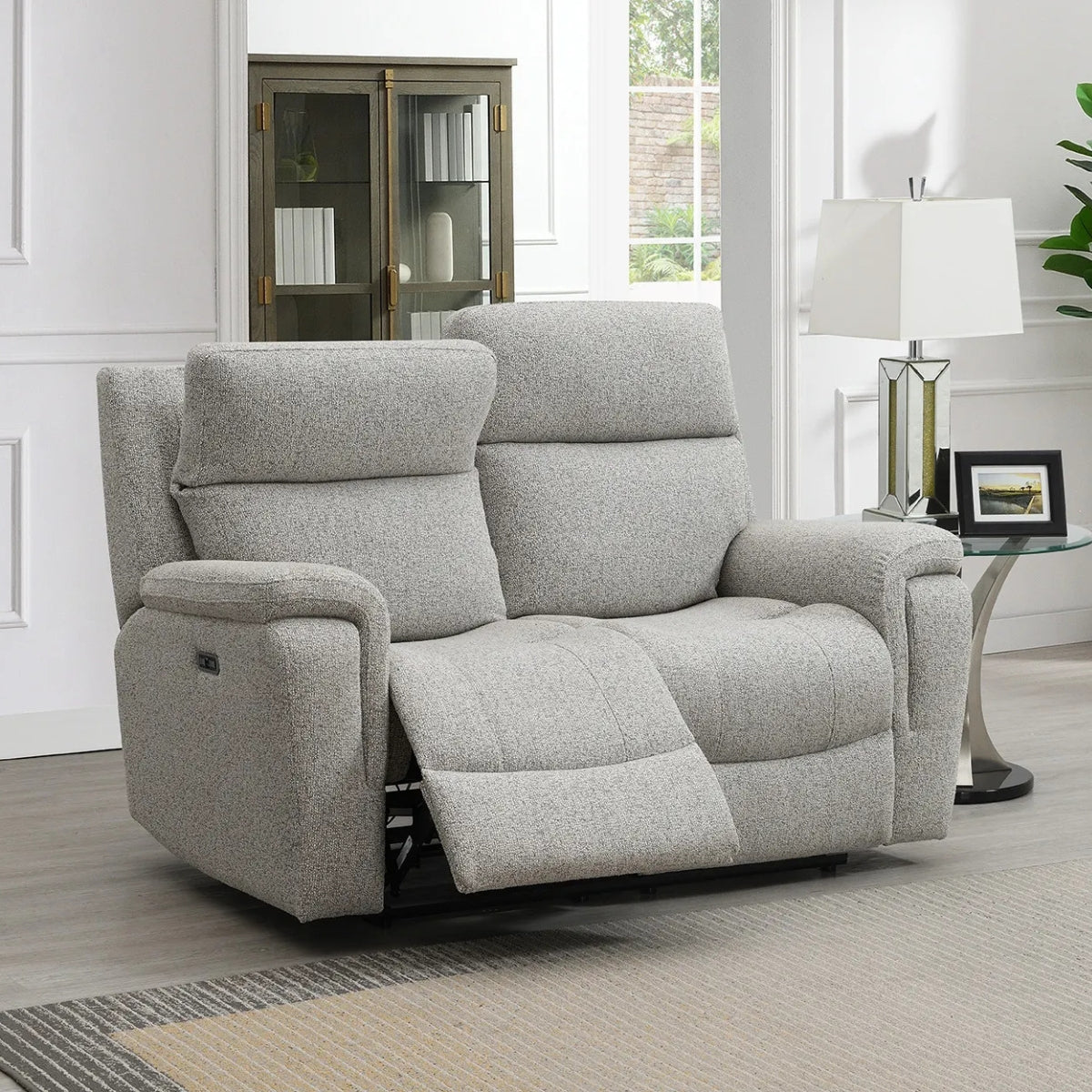 Hector grey upholstered reclining loveseat shown in a bright living room setting with a side table, lamp, window, and cabinet in the background.