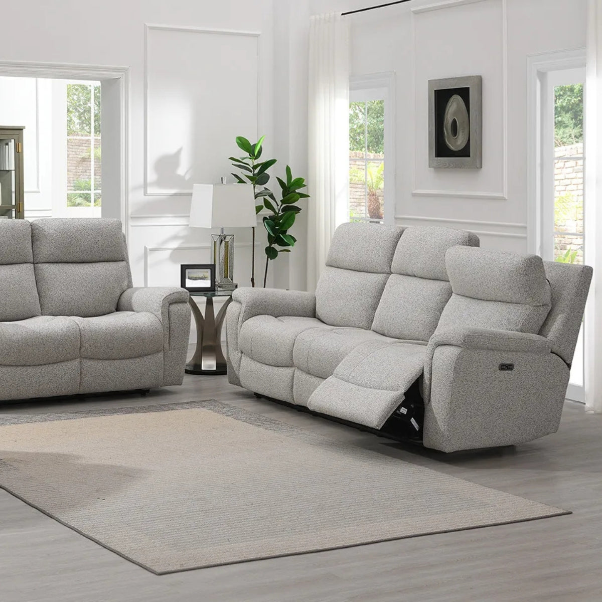 Hector grey upholstered reclining 3 seater and matching 2 seater in a bright, modern living room with white walls, large windows, and a neutral area rug.