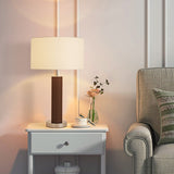 Nighstand with lamp, bowl, and vase next to a grey armchair with a patterned pillow.