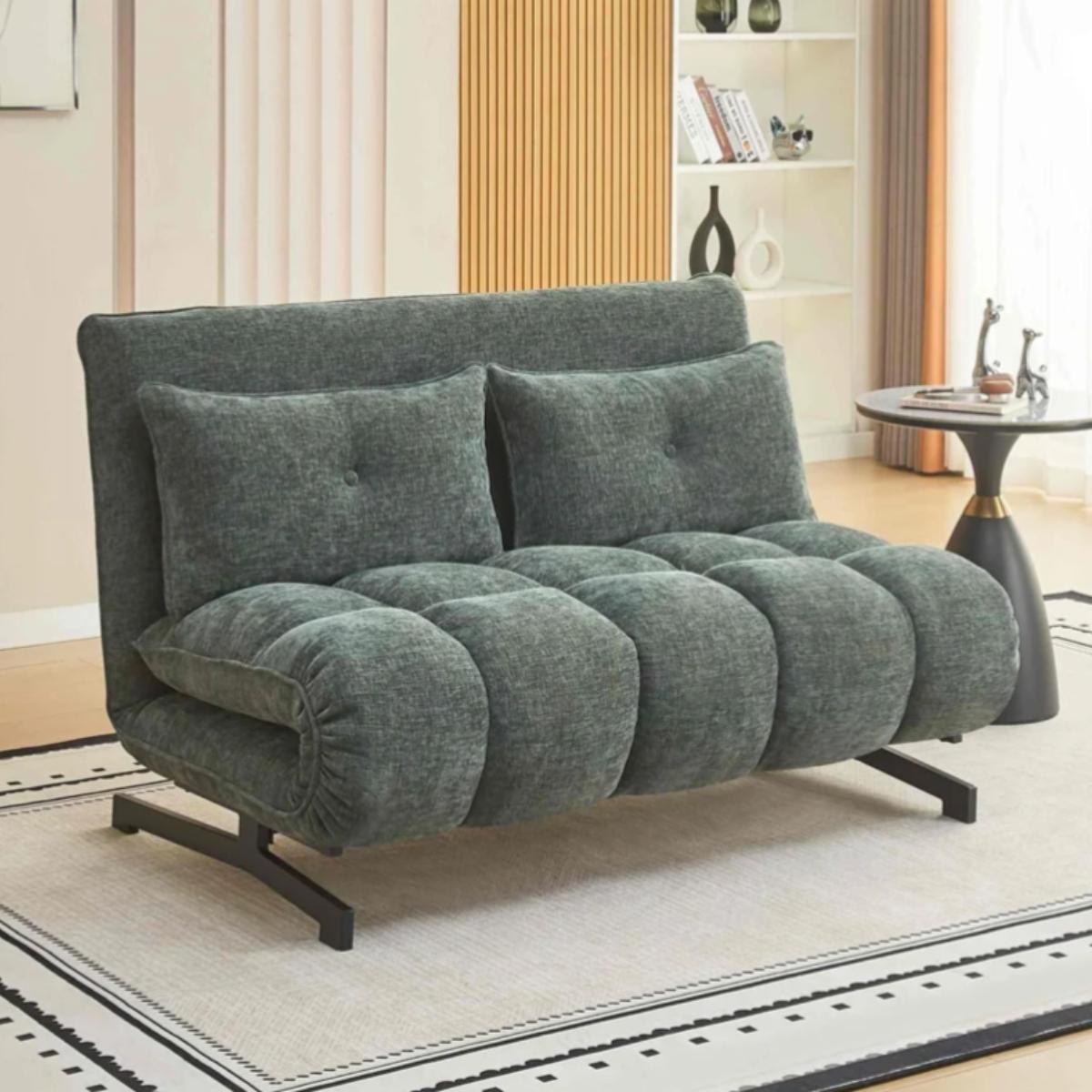 A compact upholstered futon sofa in textured dark gray fabric, styled in a modern living room setting with a patterned rug, wooden slat wall, and side table.