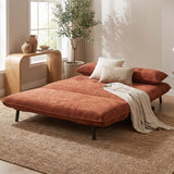 An orange-brown velvet futon daybed styled in a bright, modern room with a beige woven rug, wooden side table, potted plant, and neutral-toned pillows and throw blanket.