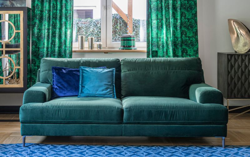 Mid century modern bottle green sofa with blue cushions, green curtain backdrop and blue rug. 
