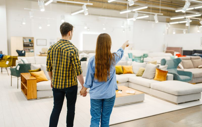 Man and woman holding hands sofa shopping with a view of cream sofas and coloured cushions. 